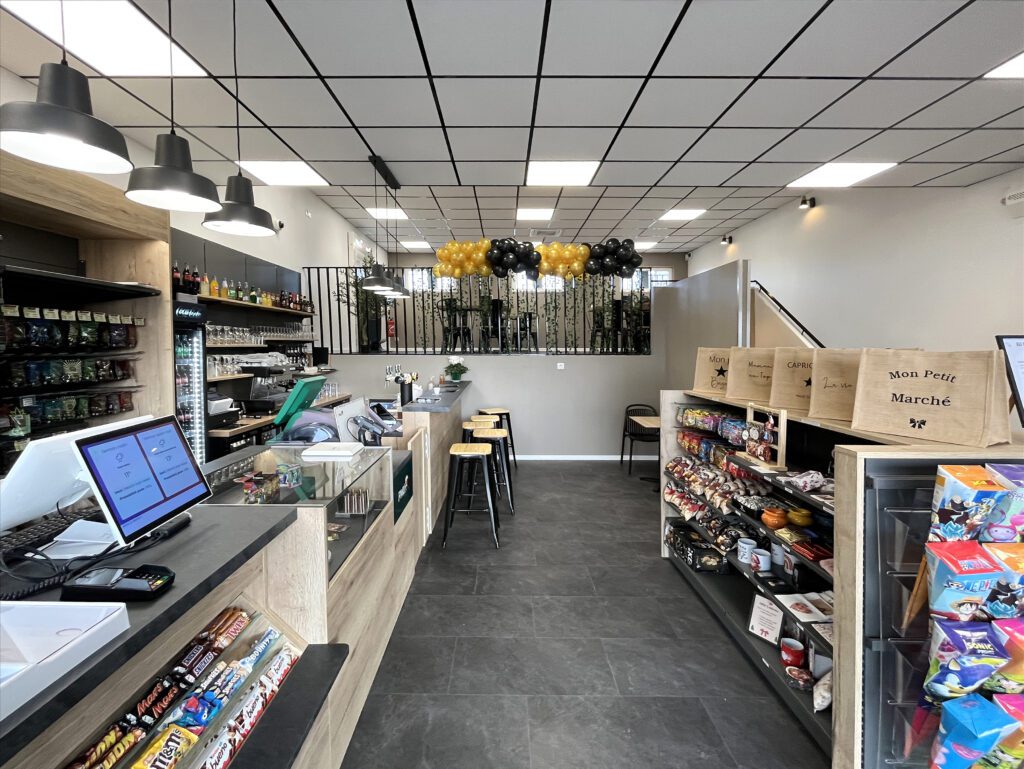 Agencement moderne de bar tabac presse avec comptoir en bois clair, espace caisse, étagères de confiseries et bar avec tabourets sous luminaires suspendus.