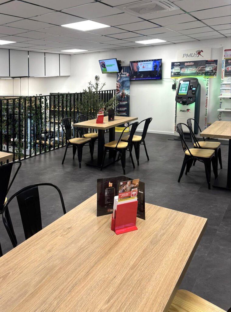 Espace bar convivial avec tables en bois, chaises noires, écrans muraux et borne PMU dans un environnement lumineux et contemporain.
