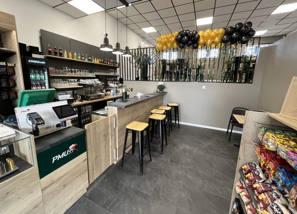 Espace bar et comptoir PMU avec tireuse à bière, tabourets hauts et décoration sobre mêlant bois, métal et touches végétales.
